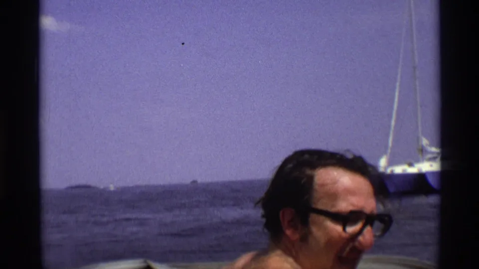 A vintage New York river frame shows an adult man wearing dark glasses in the foreground, with open water and a sailboat behind him.