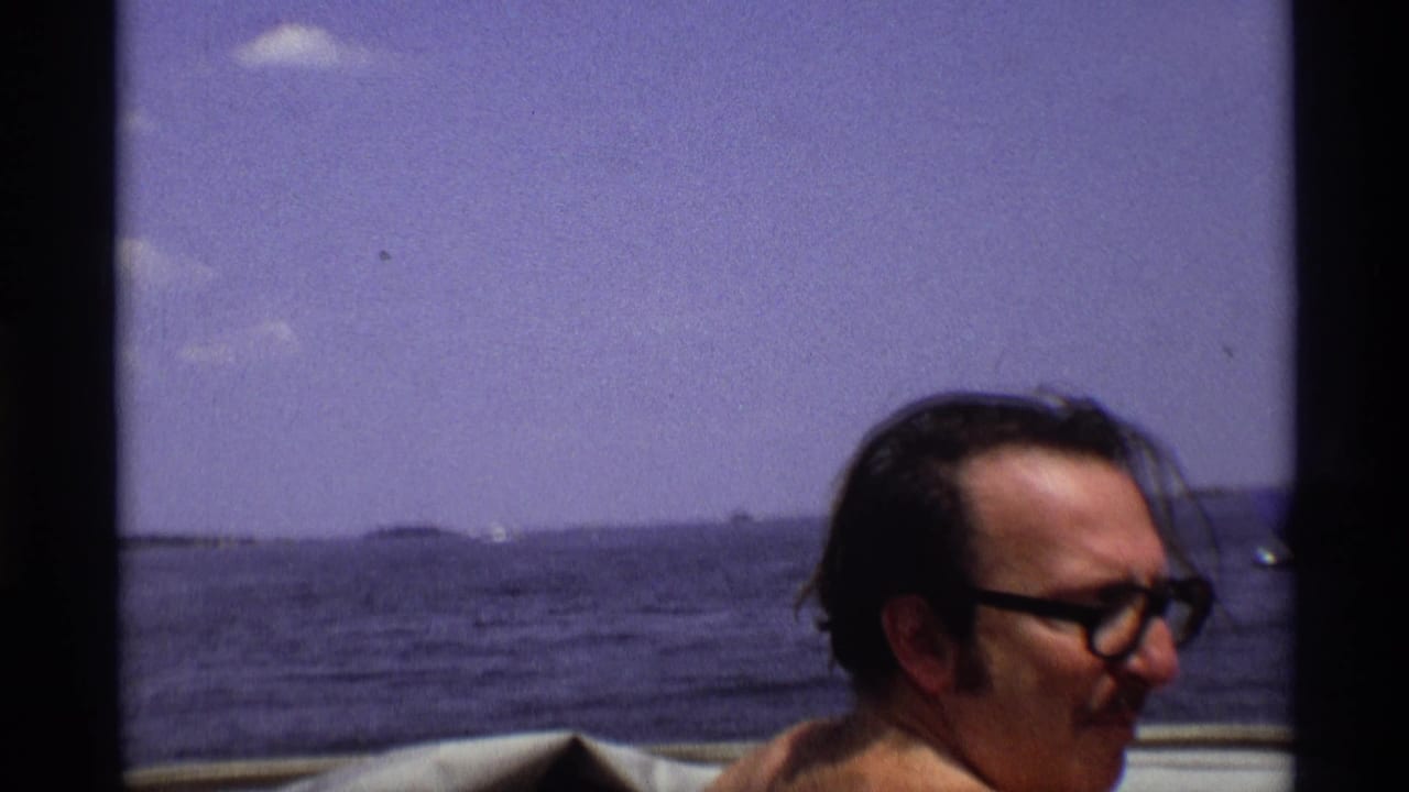 Man in glasses seated on a boat with open water and distant land behind him.