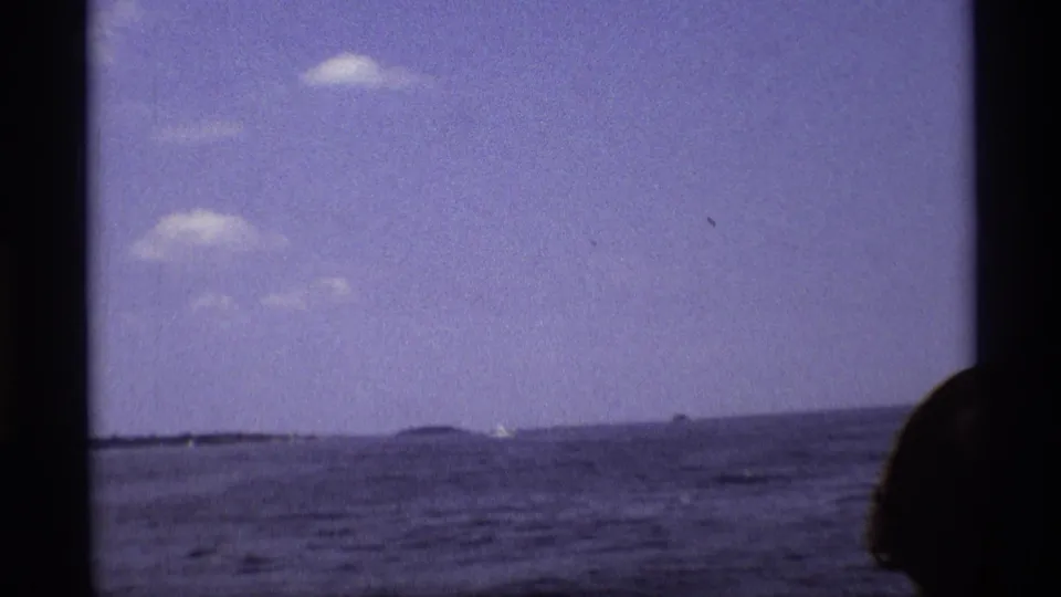 1973 New York 8mm frame looking across open water from a boat, with distant land and a partial passenger silhouette at the right edge.