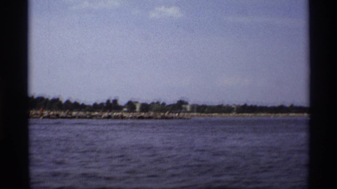 1973 New York 8mm frame of water and a distant shoreline, with dark boat framing dividing the home-movie view.