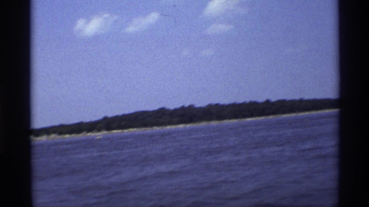 1973 New York 8mm frame showing blue lake water, a distant tree-lined shore, and scattered clouds from a moving boat viewpoint.