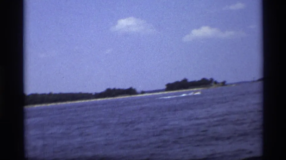 1973 New York 8mm frame with a small speedboat moving across blue water near a low wooded shoreline.
