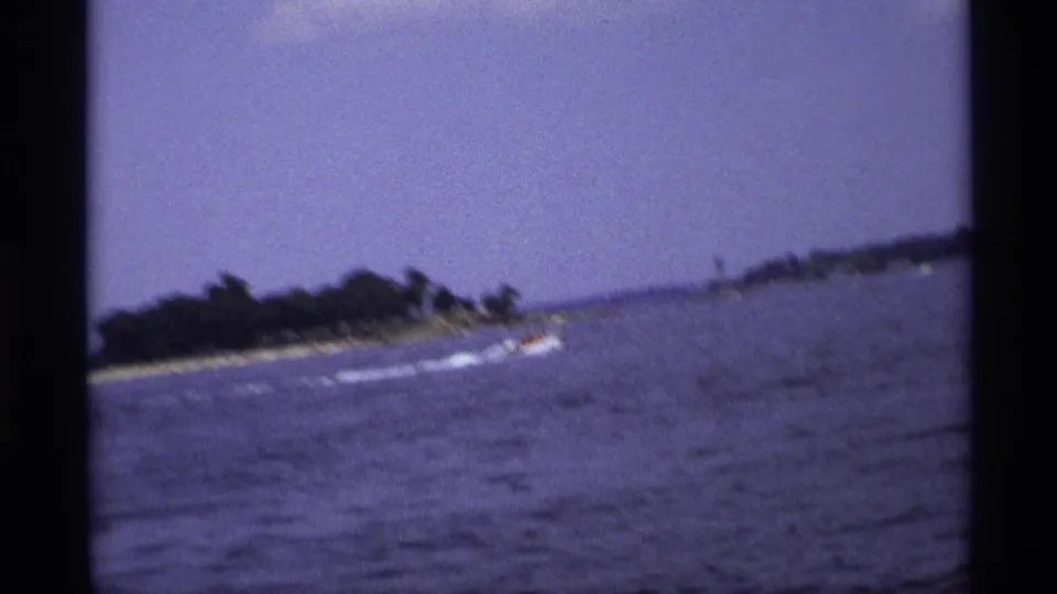 1973 New York 8mm frame of a small speedboat gliding by a wooded point on blue lake water.