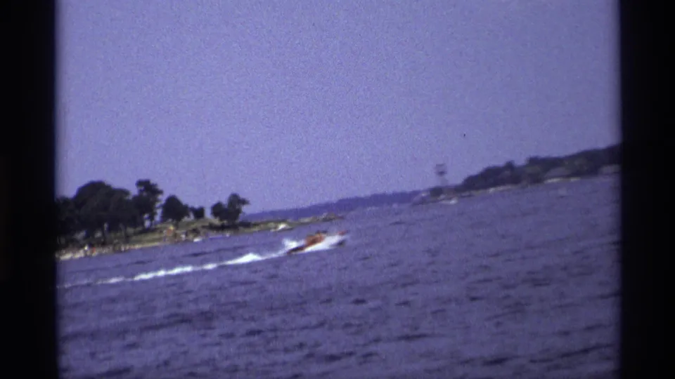 1973 New York home-movie frame showing a small motorboat crossing open water with a white wake near a low, tree-lined shoreline.