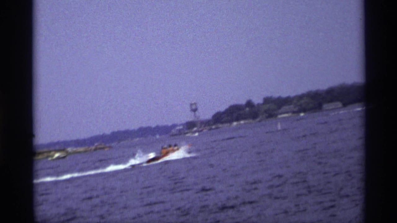 Motorboat on open water with wake and distant shoreline behind it.
