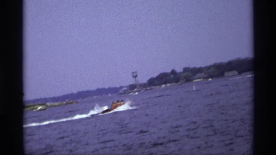 A small motorboat travels across open water in the 1973 New York home-movie footage, with shoreline and a distant tower-like structure visible.
