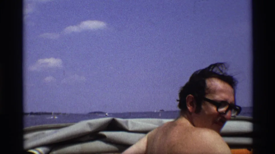 A shirtless adult man wearing glasses sits in a boat during the 1973 New York footage, with windblown hair and open water behind him.