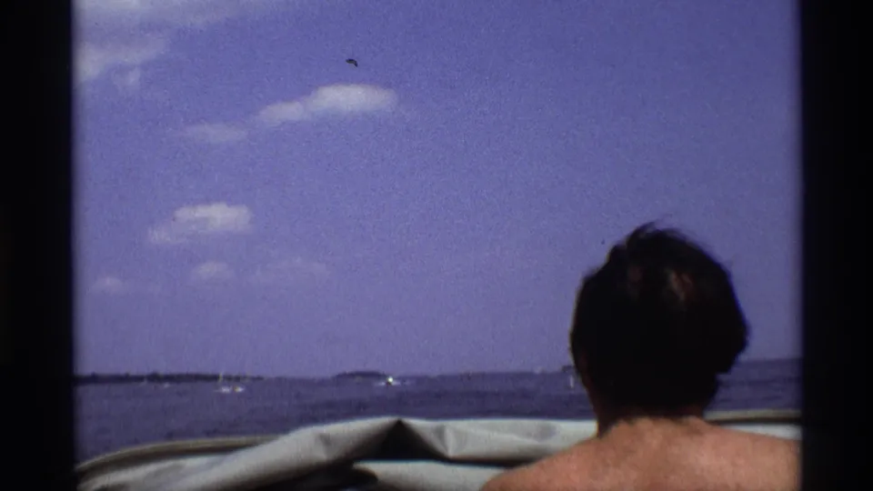 The final frame shows the back of a shirtless adult man in a boat, facing open water under a blue sky in the 1973 New York home movie.