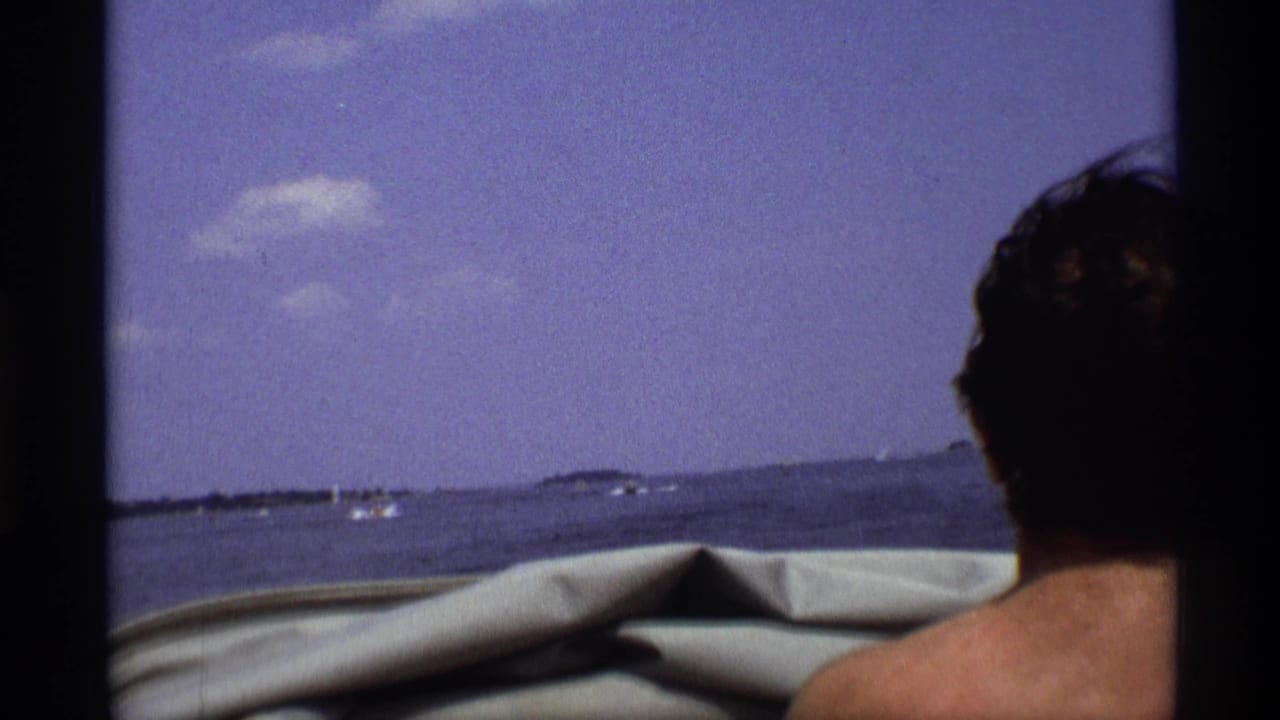 Man at right edge of a boat frame with lake and small boats ahead.