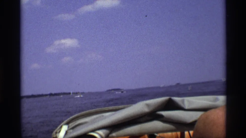 1973 New York home-movie frame showing the boat's front edge, open lake, distant shoreline, and part of a passenger at right.
