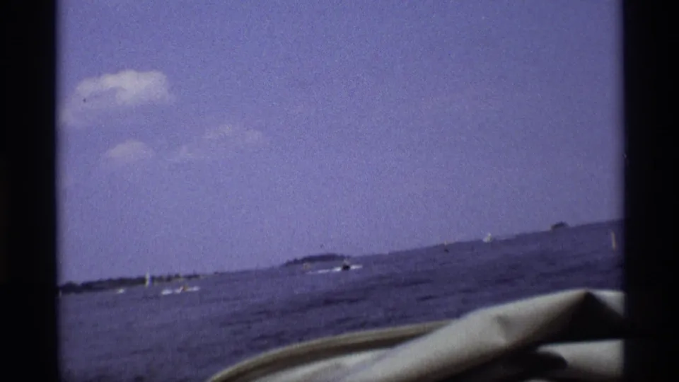 1973 New York 8mm frame with a slightly tilted boat-level view over lake water, distant boats, shoreline, and pale sky.
