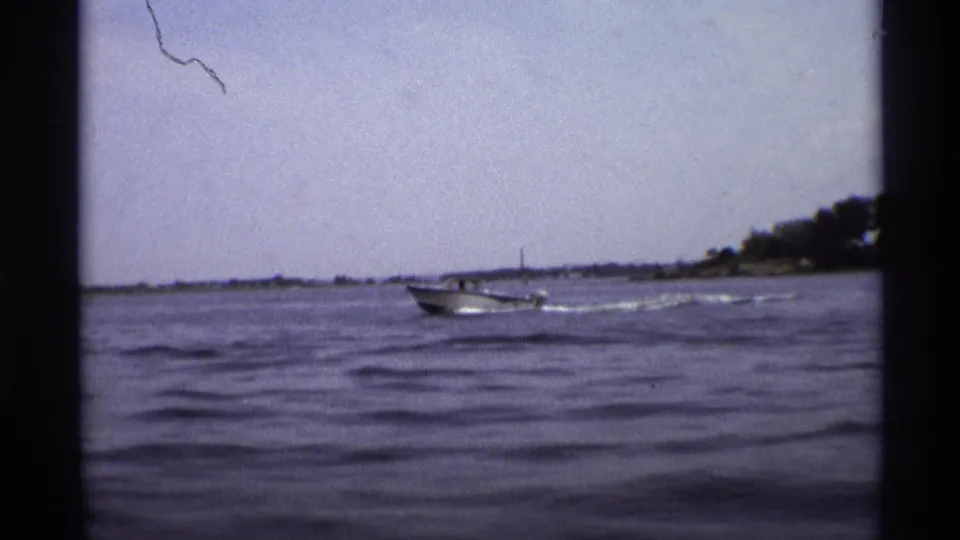 A motorboat travels along a New York lake near a low shoreline, seen through a soft, grainy 8mm frame.