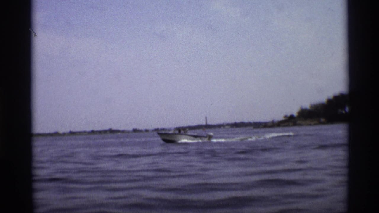 1973 New York 8mm frame showing a small motorboat moving across wavy water, with shoreline and a distant tower on the horizon.