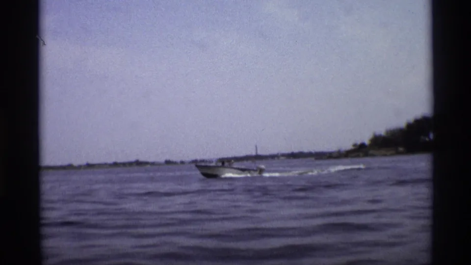 1973 New York 8mm frame showing a small motorboat moving across wavy water, with shoreline and a distant tower on the horizon.