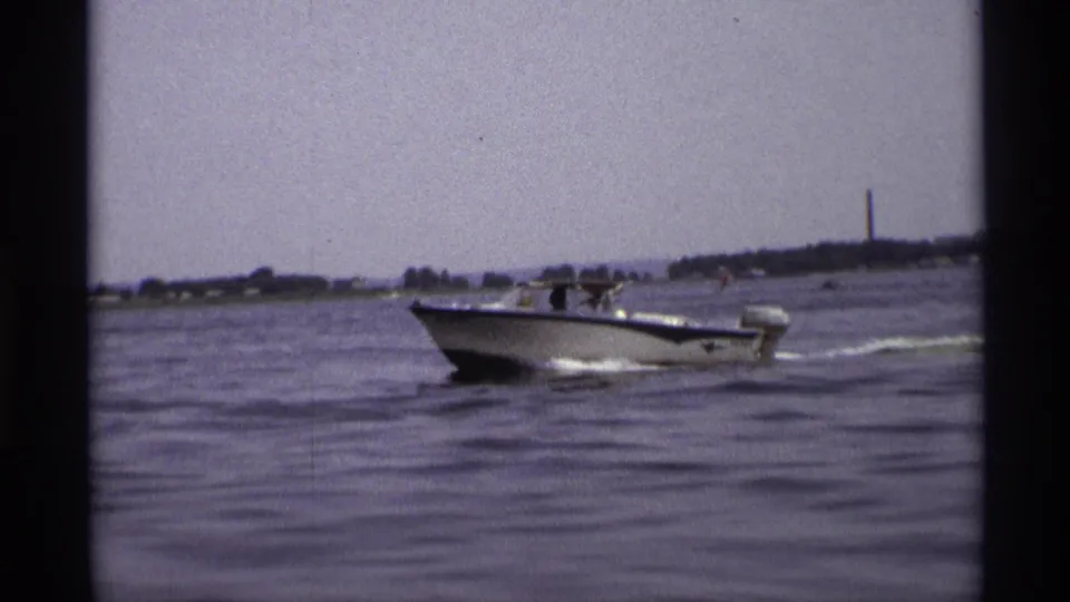 1973 New York 8mm frame showing a closer small motorboat with people aboard, crossing wavy water near a low shoreline.