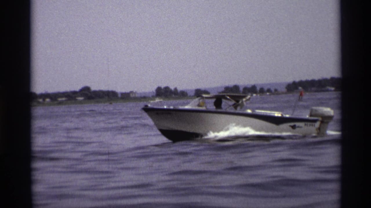 1973 New York 8mm frame of a white and blue motorboat moving across a lake with people aboard and a distant shoreline.