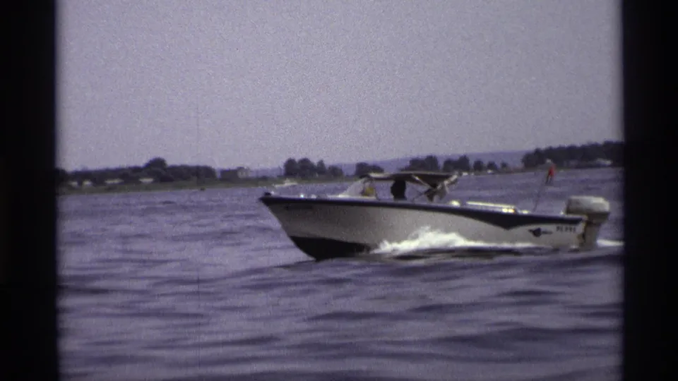 1973 New York 8mm frame of a white and blue motorboat moving across a lake with people aboard and a distant shoreline.
