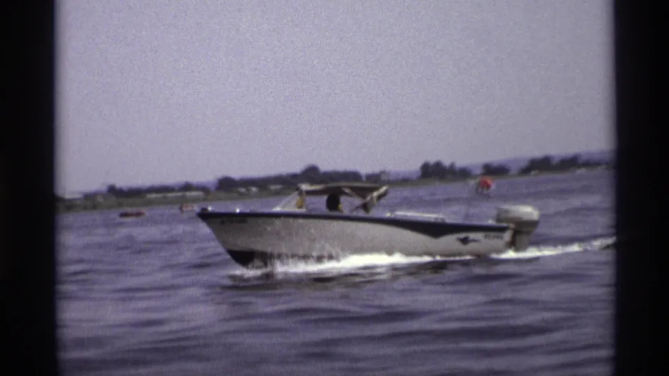 The motorboat continues across the lake in a 1973 New York home-movie frame, with rippled water and a low tree-lined shore behind it.