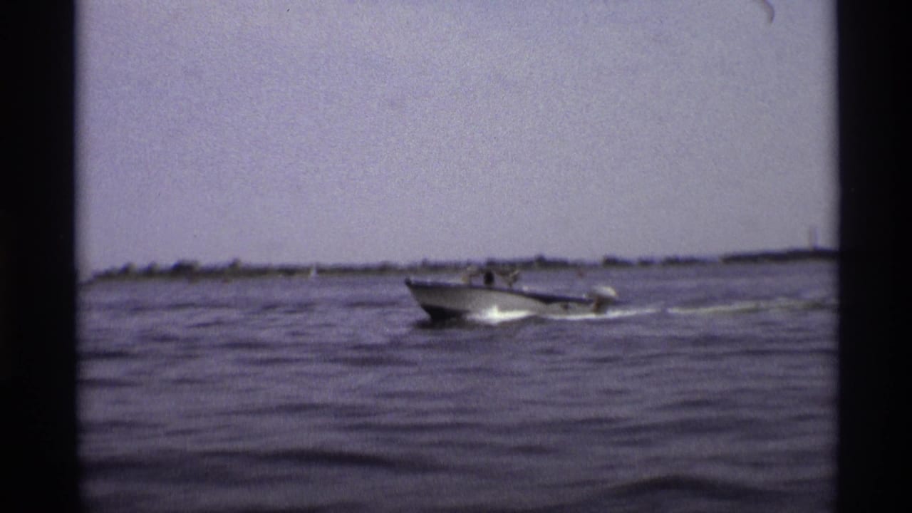 A 1973 New York home-movie frame shows the motorboat at mid-distance on rippled water under a hazy sky.