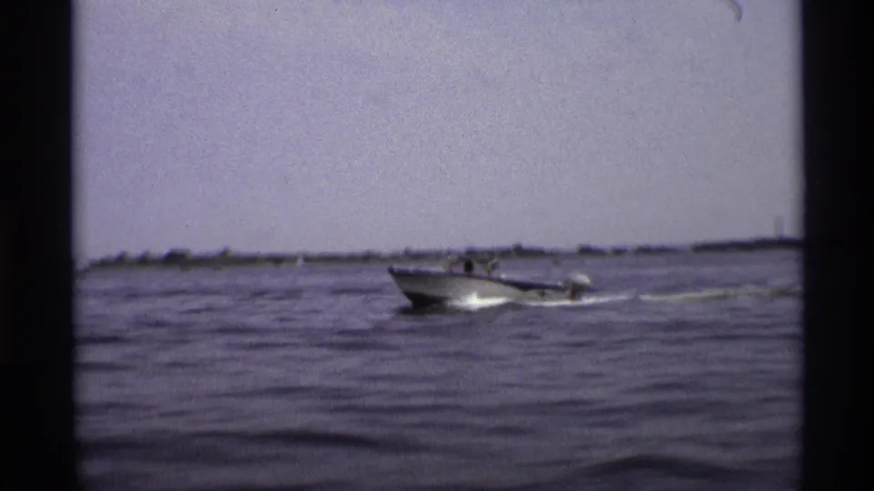 A 1973 New York home-movie frame shows the motorboat at mid-distance on rippled water under a hazy sky.