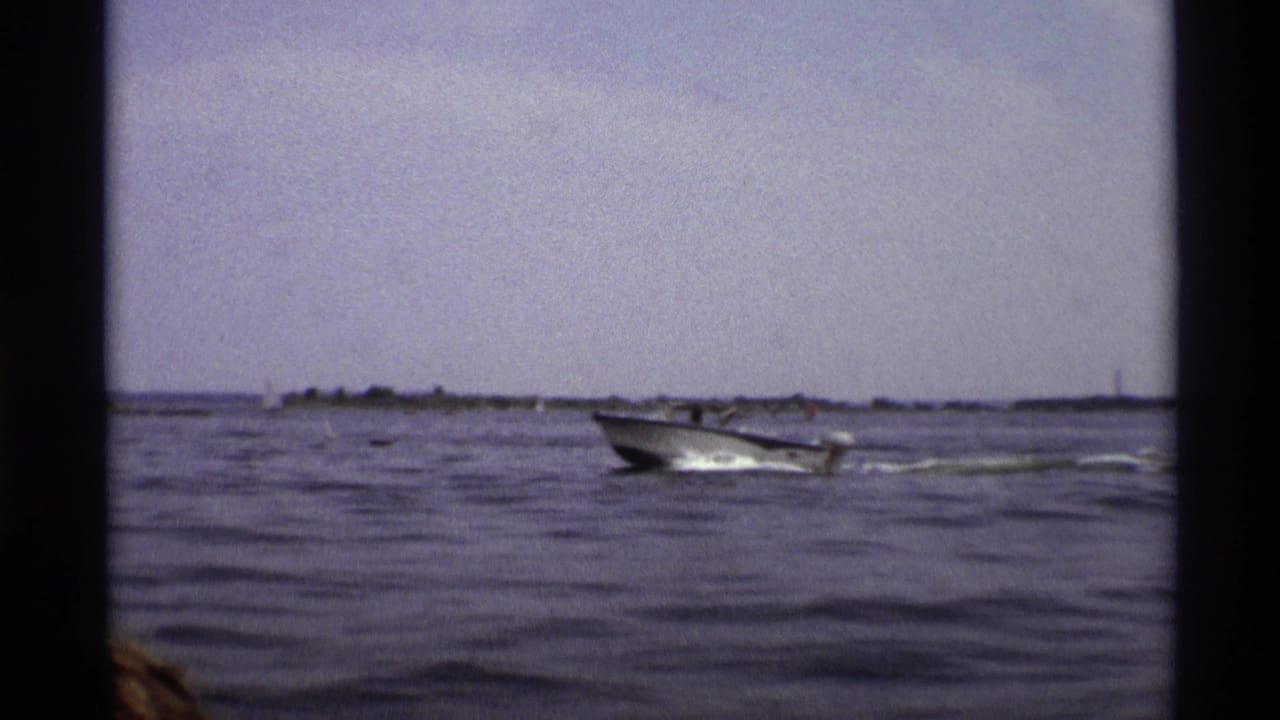 The blue and white motorboat passes through a wide 1973 New York water view with distant shoreline and small sailboats visible.