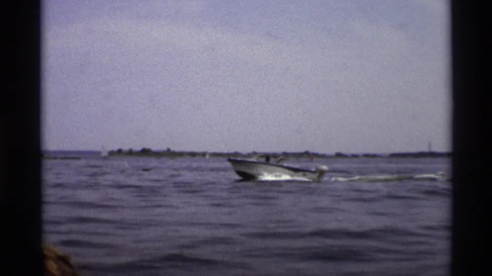 The blue and white motorboat passes through a wide 1973 New York water view with distant shoreline and small sailboats visible.