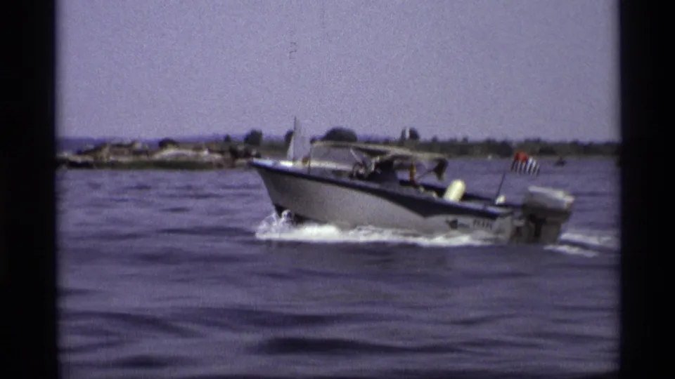 A closer 1973 New York frame shows the motorboat passing with a canopy, outboard motor, wake, and small American flag at the stern.