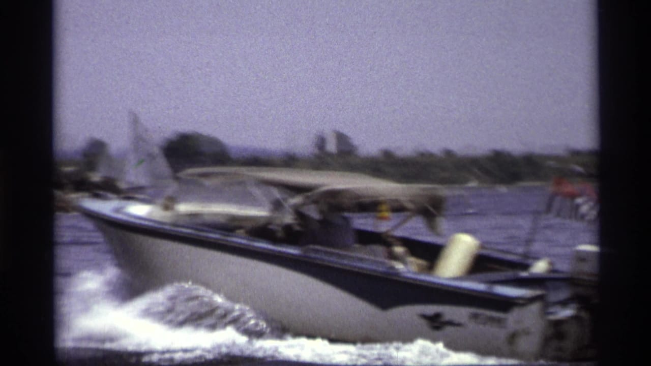 The motorboat passes close to the camera on New York water, with spray, wake, canopy, flag, and shoreline softened by motion.