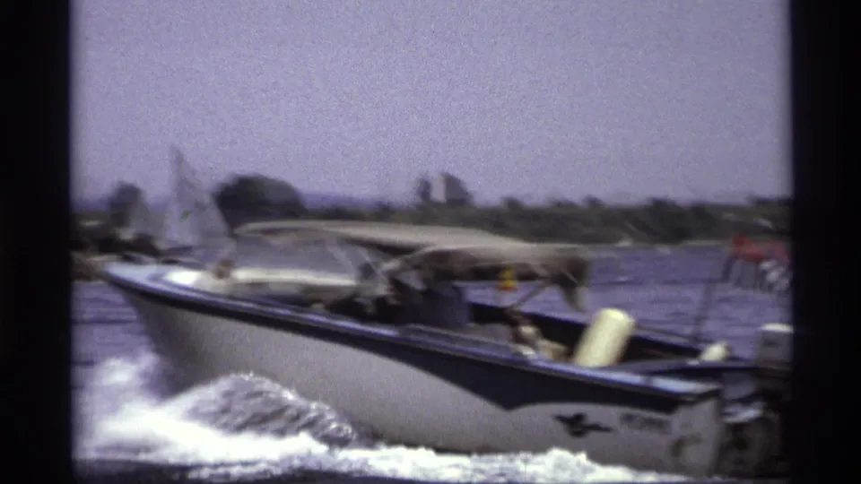 The motorboat passes close to the camera on New York water, with spray, wake, canopy, flag, and shoreline softened by motion.