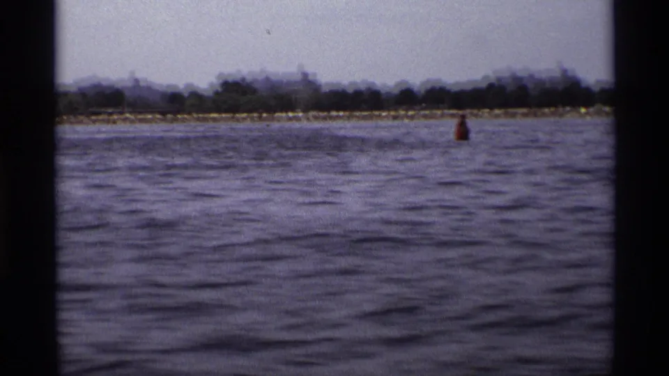 1973 New York 8mm frame showing water from a moving boat, a distant wooded shore, low buildings, and a red buoy.