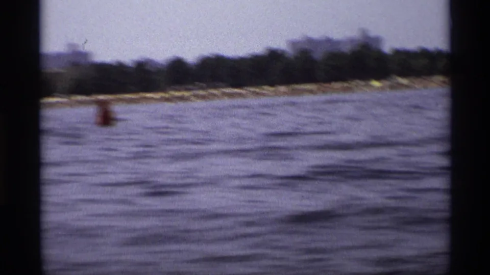Soft 8mm view from a boat across New York water toward a wooded shoreline, low structures, and a red buoy.