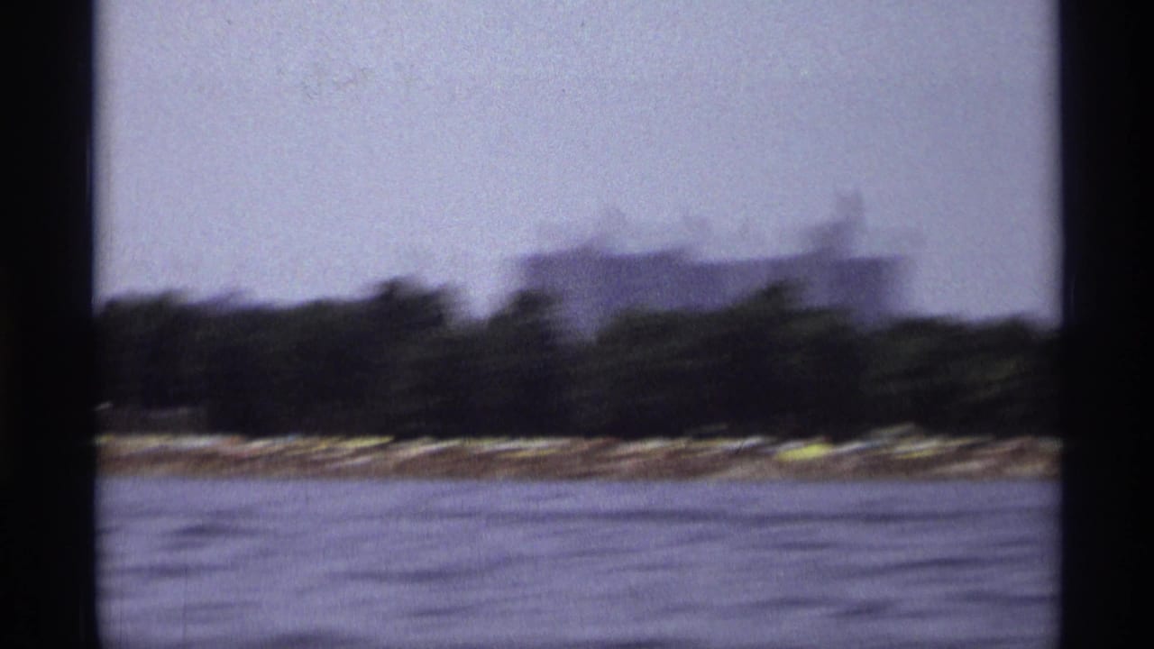 Moving 1973 New York boat footage with blurred water, shoreline trees, and a low building in the distance.
