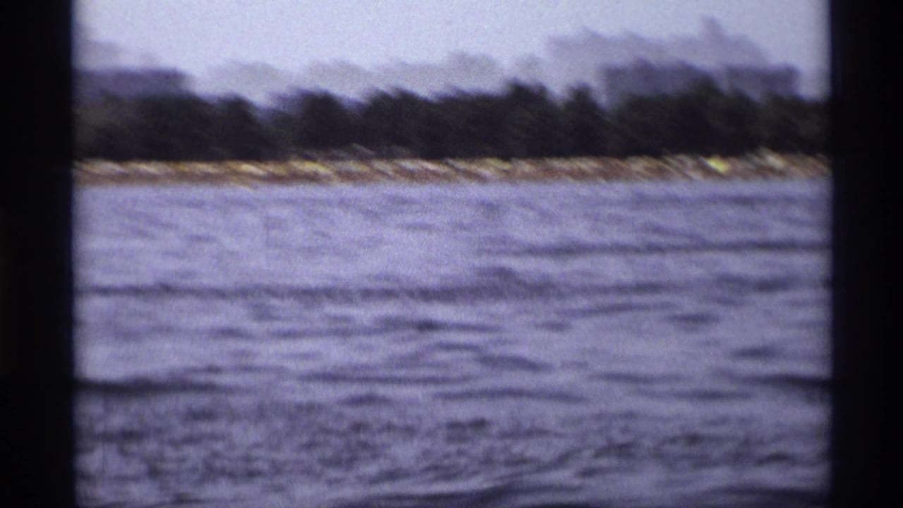 Faded 8mm frame of a New York shoreline seen from a moving boat, with water, trees, and distant low buildings.