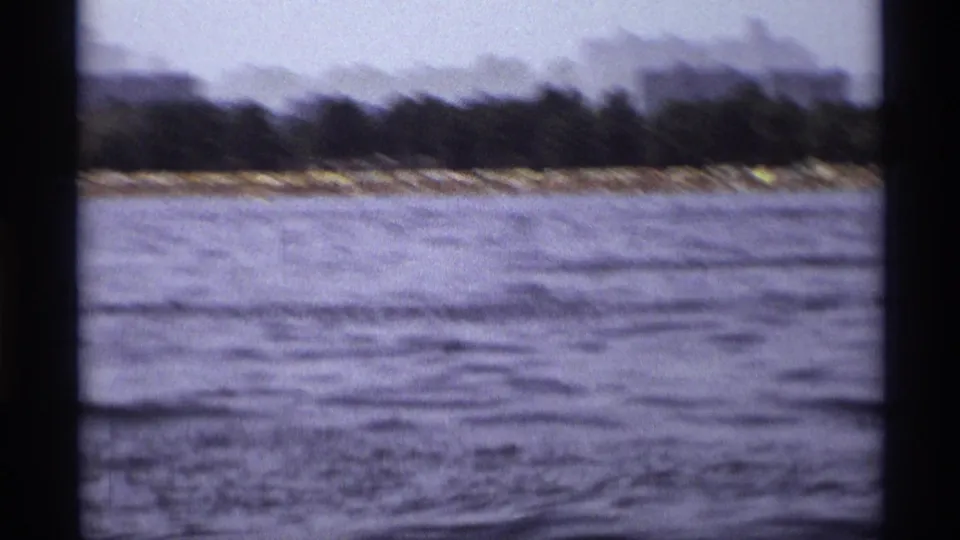 Faded 8mm frame of a New York shoreline seen from a moving boat, with water, trees, and distant low buildings.