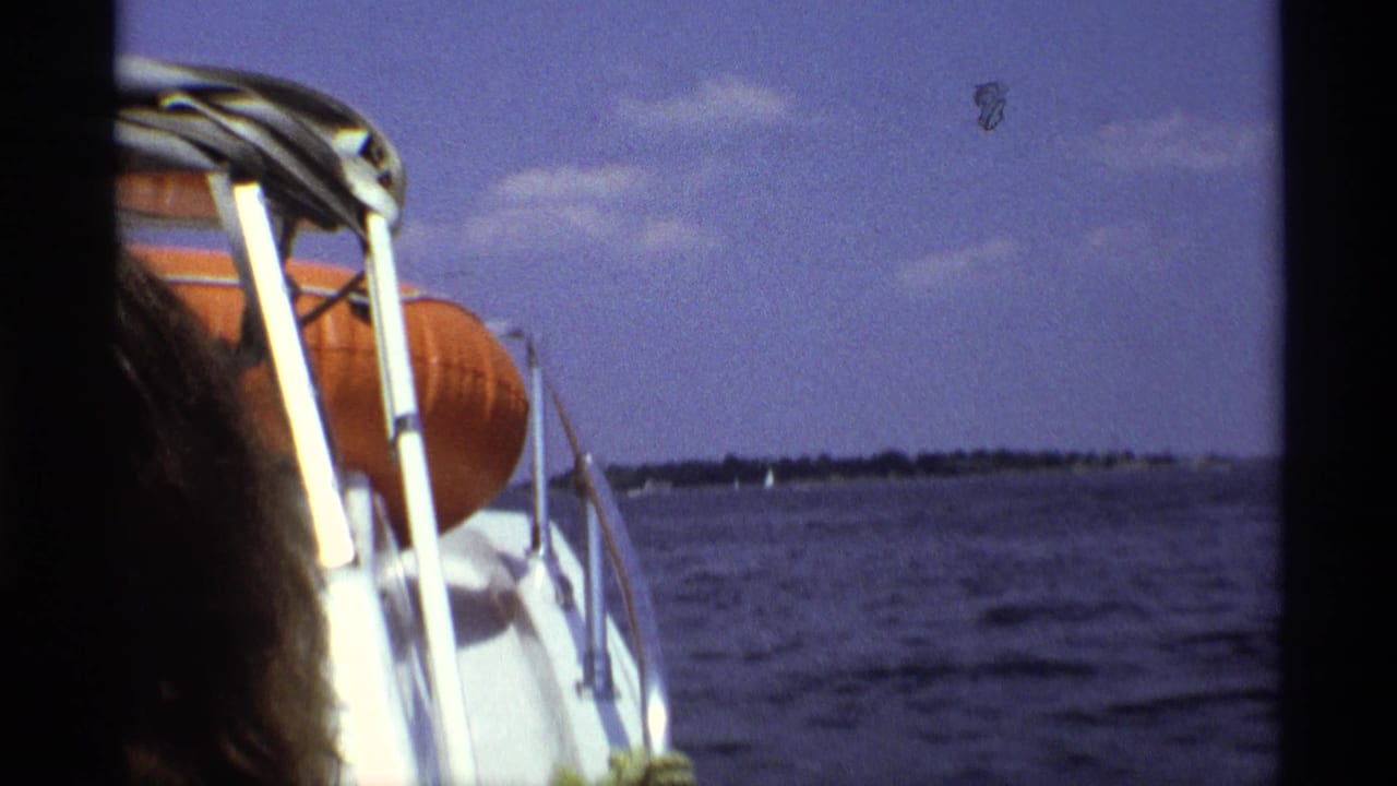 1973 New York 8mm boating frame with a partial passenger, white railings, orange life preserver, and distant shoreline across the water.