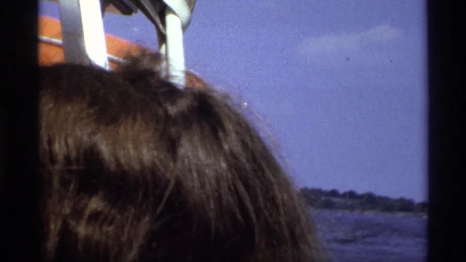 1973 New York 8mm frame showing the top of a passenger's head, white boat railings, water, and a distant tree-lined shore.