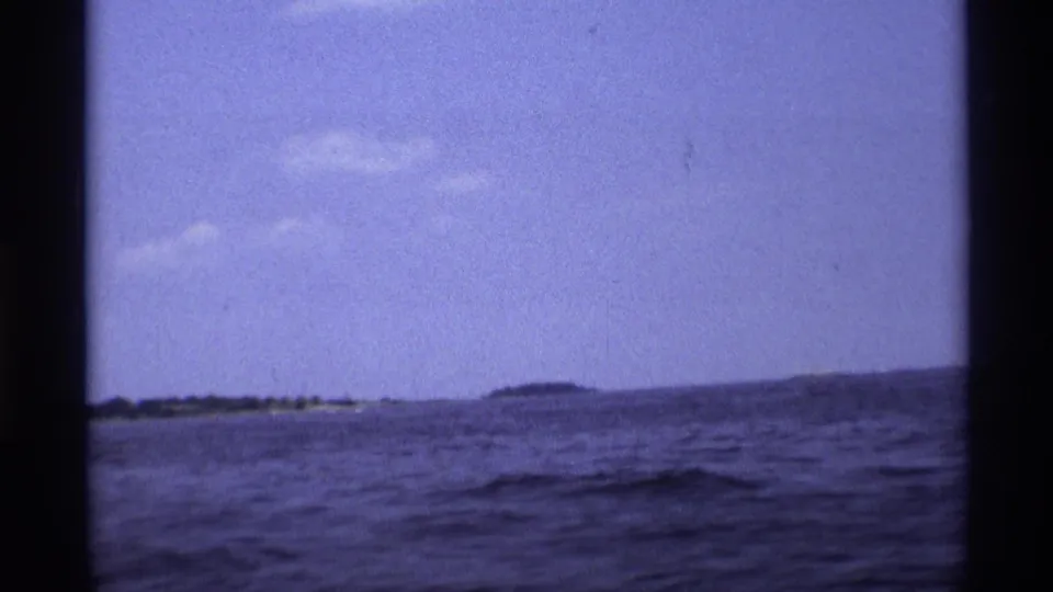1973 New York 8mm frame showing open water, a low distant shoreline, blue sky, and the dark edges of the boat or film gate.