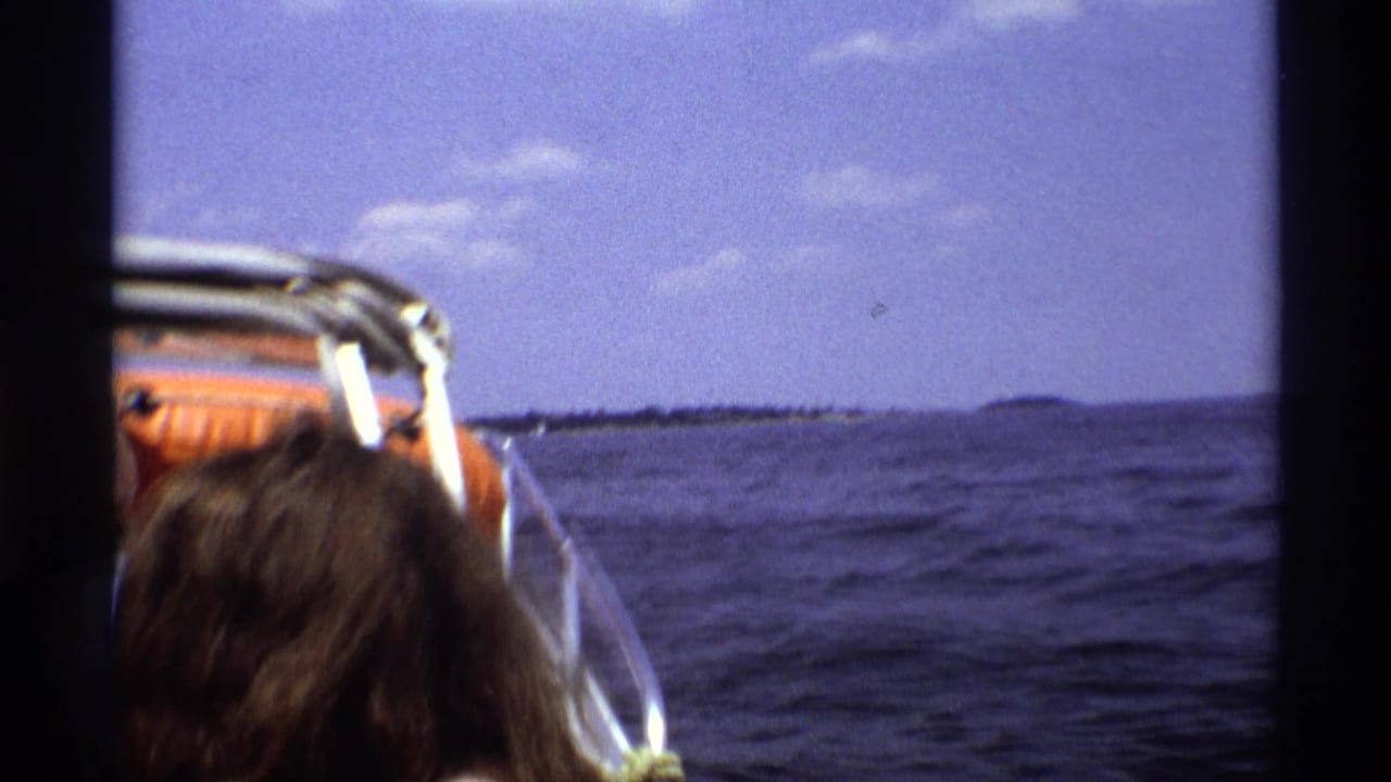 A 1973 New York boat-ride frame with a passenger seen from behind, an orange seat or cushion, rippled water, and a distant shoreline.