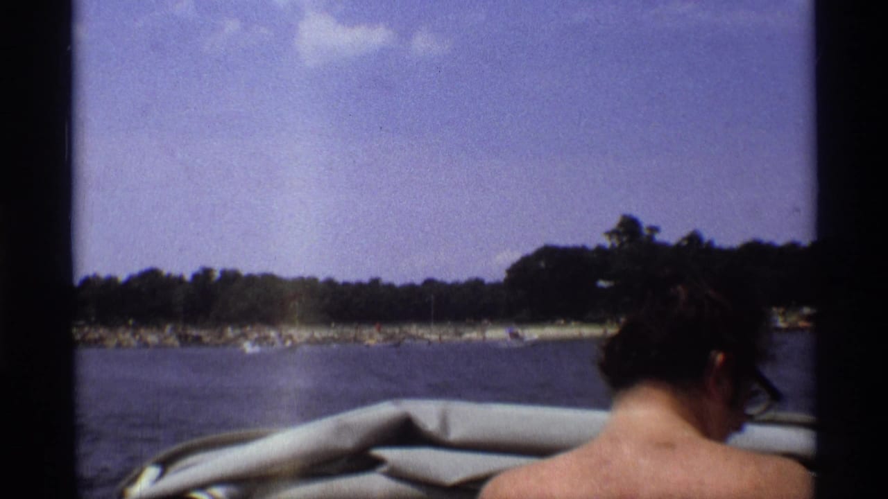 The boat nears a wooded New York shoreline, with a passenger seen from behind, pale boat upholstery, water, trees, and sky visible.
