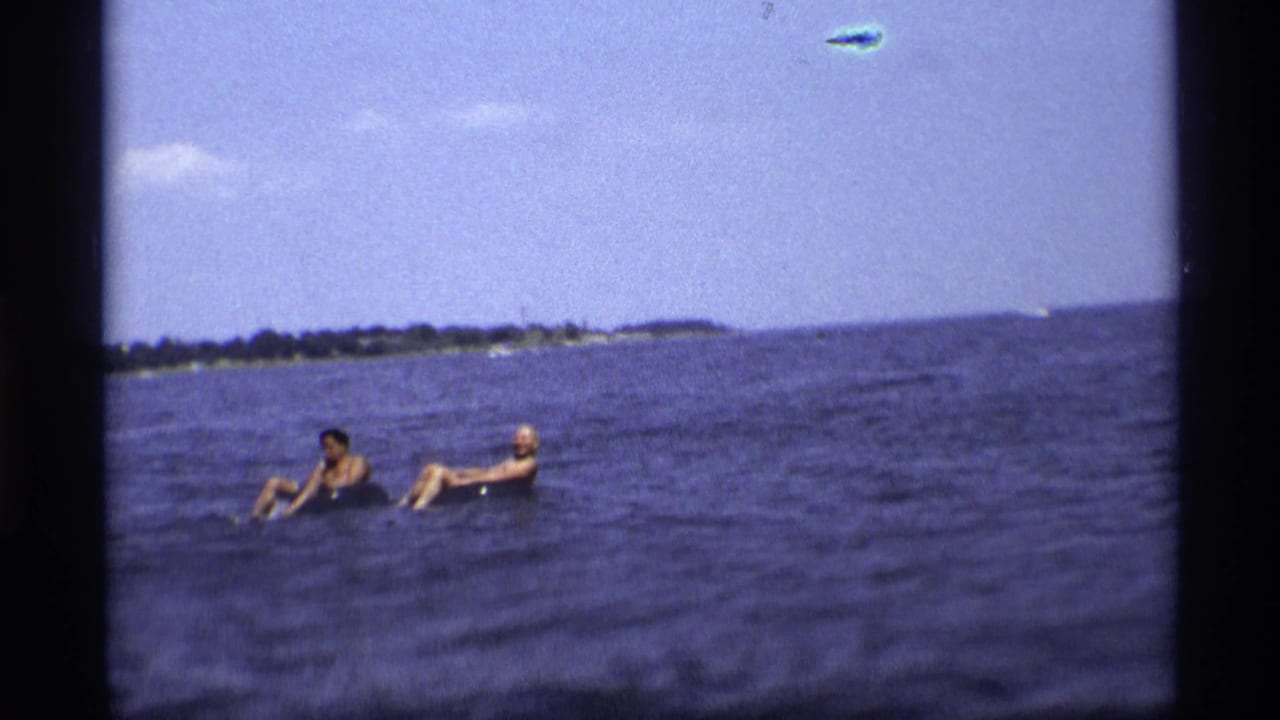 Two people float in inflatable tubes on open lake water near a distant shoreline.