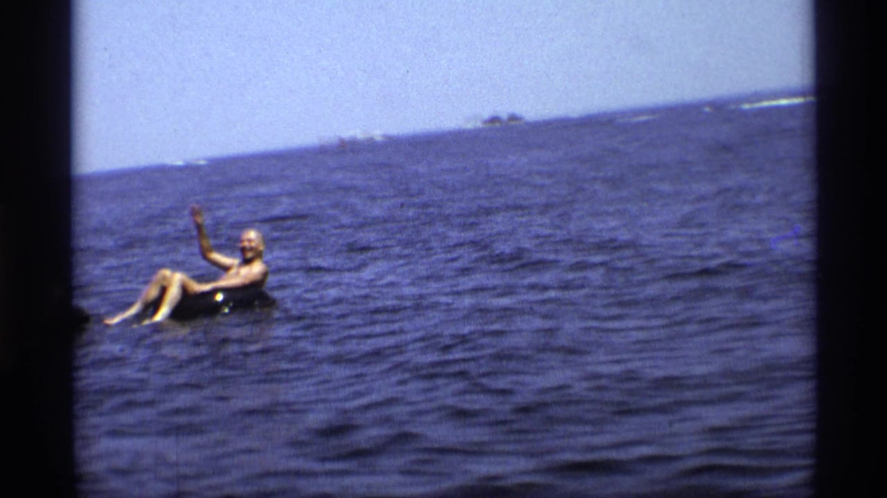 A man in a dark swimsuit waves while floating in an inflatable tube on the lake.