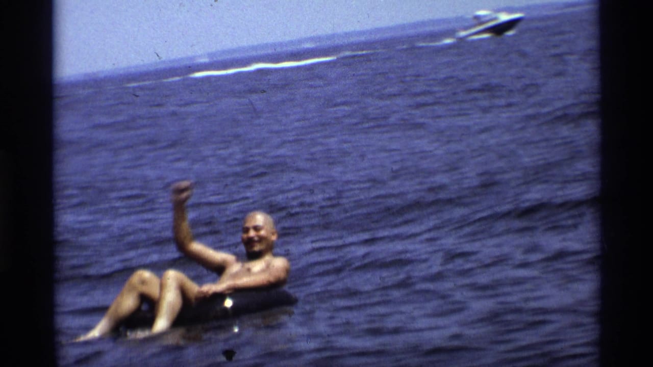 Closer 8mm frame of a man smiling and waving from a black tube on a New York lake in 1973.