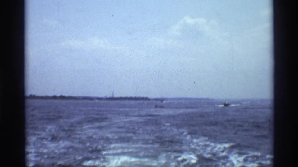 Wide 1973 New York lake view with wake trailing across the water and small boats near the distant shore.