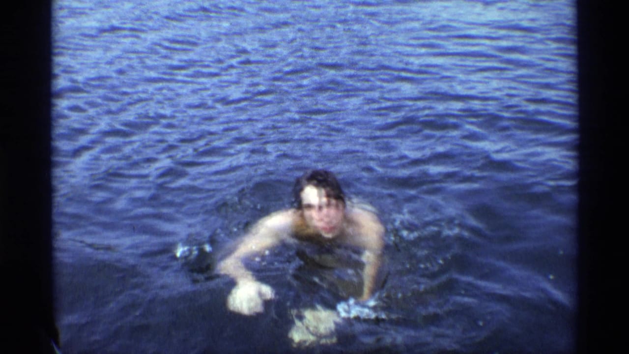 The swimmer faces the camera in blue water, with his shoulders and arms breaking the surface.