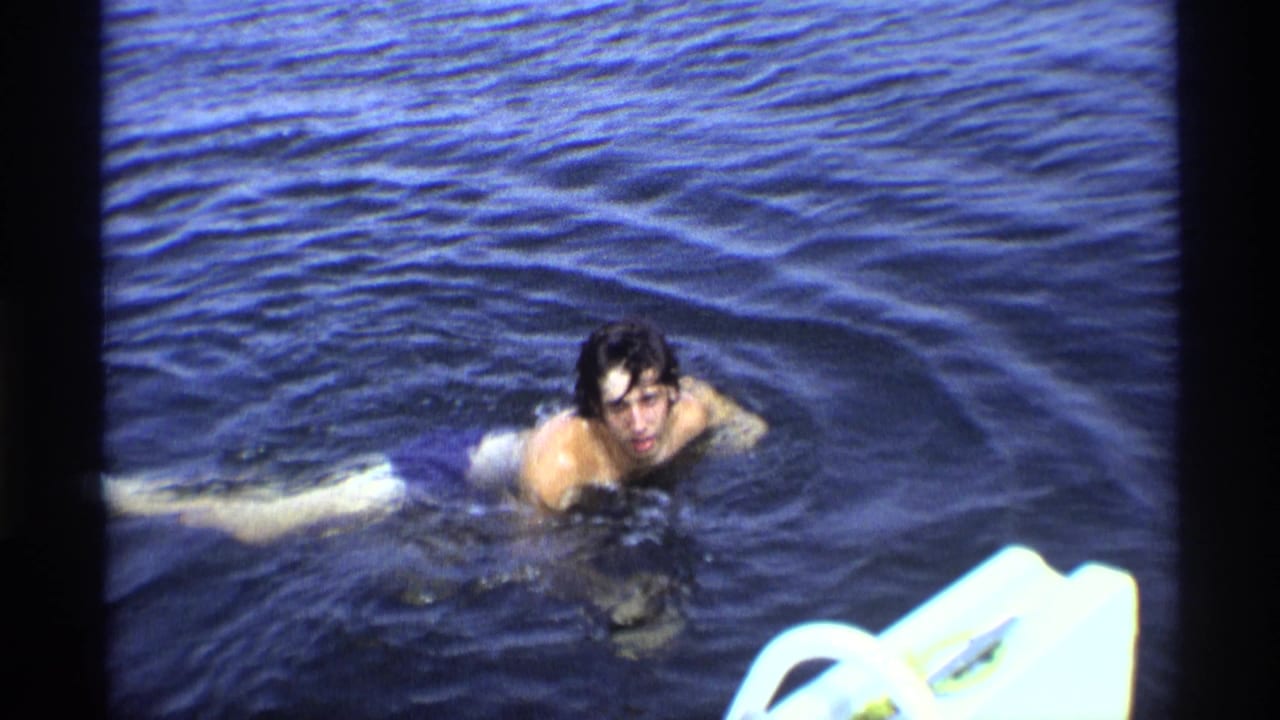 In soft 8mm color, the swimmer approaches through rippling water beside a bright white object at frame edge.
