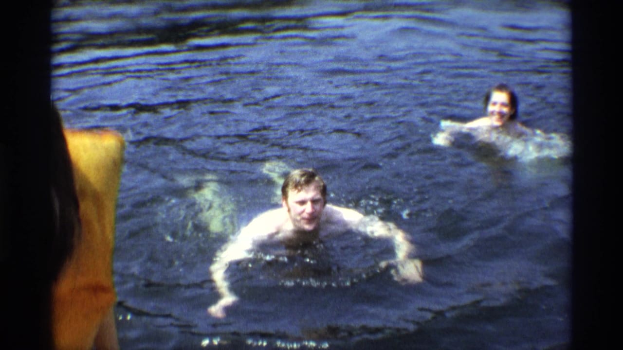 1973 New York 8mm frame showing two adults swimming in a lake, with one swimmer facing the camera and a partial figure at the edge.
