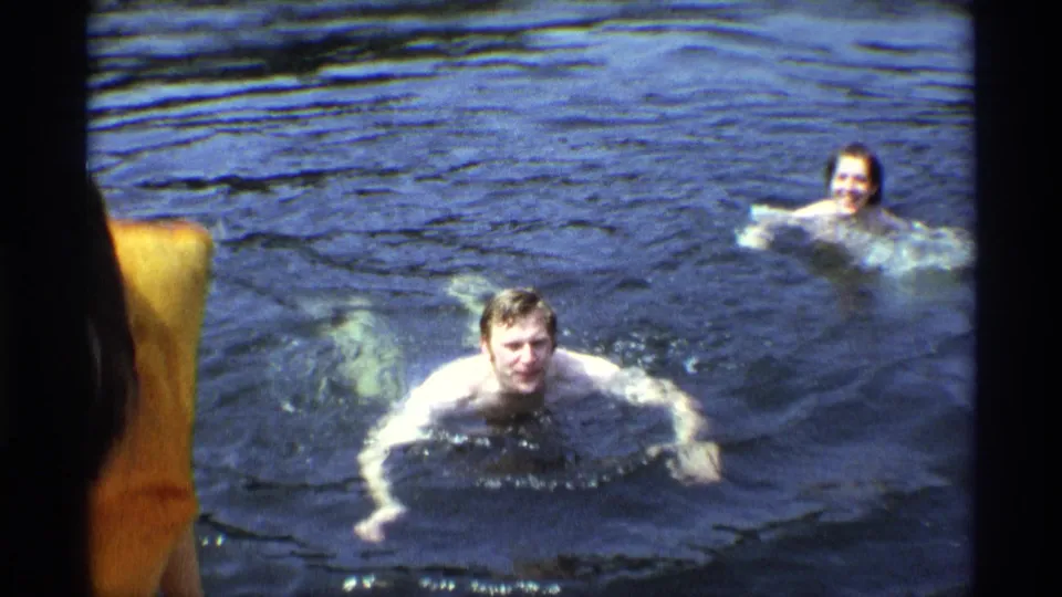 1973 New York 8mm frame showing two adults swimming in a lake, with one swimmer facing the camera and a partial figure at the edge.