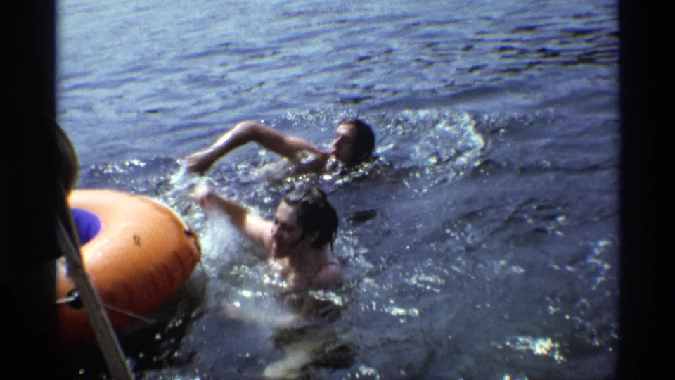 1973 New York home-movie frame with two adults swimming near an orange inflatable tube, filmed from a close lakeside or boat angle.