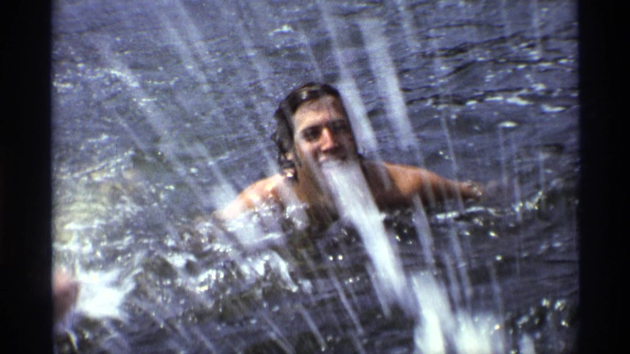 Long-haired man swimming in open water while splashing toward the camera.