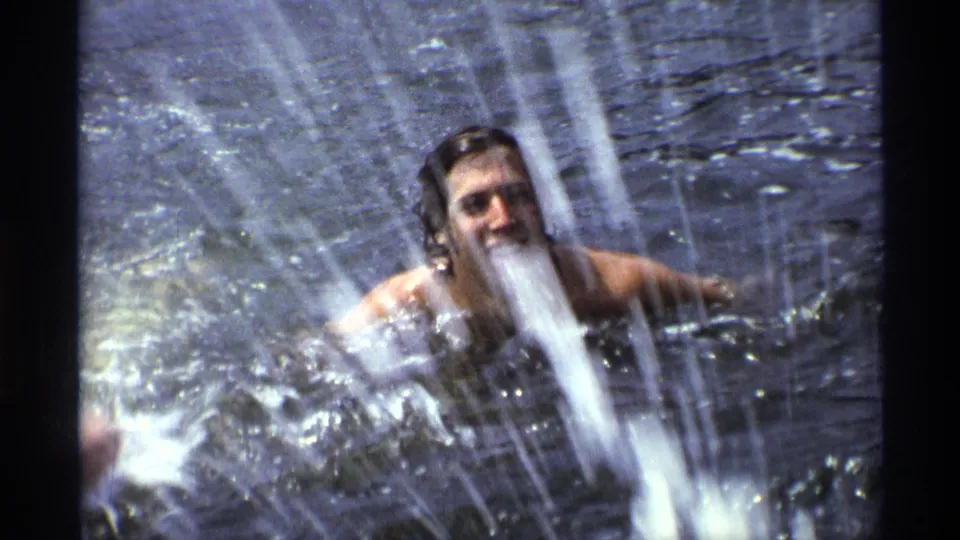 In 1973 New York home-movie footage, a long-haired man swims in open water and splashes with his arms raised.